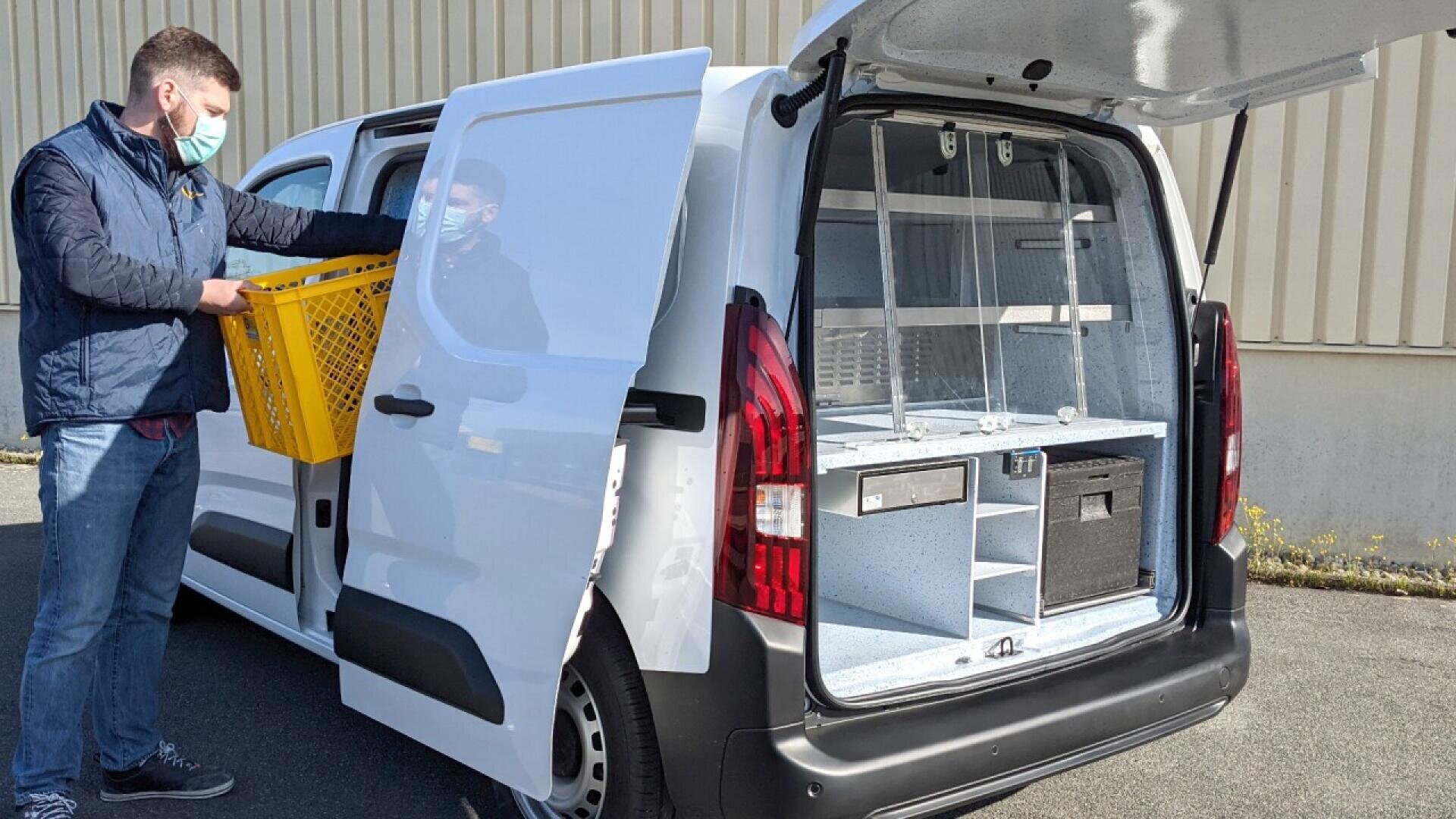 A man wearing a mask and blue jacket loads a yellow crate into the open side door of a white delivery van. The van’s rear hatch is also open, revealing organized storage compartments inside.