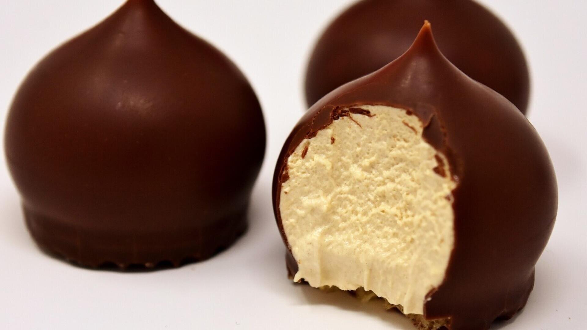 Three chocolate-covered marshmallow treats are shown on a white surface, with one treat bitten to reveal a fluffy, creamy filling inside.