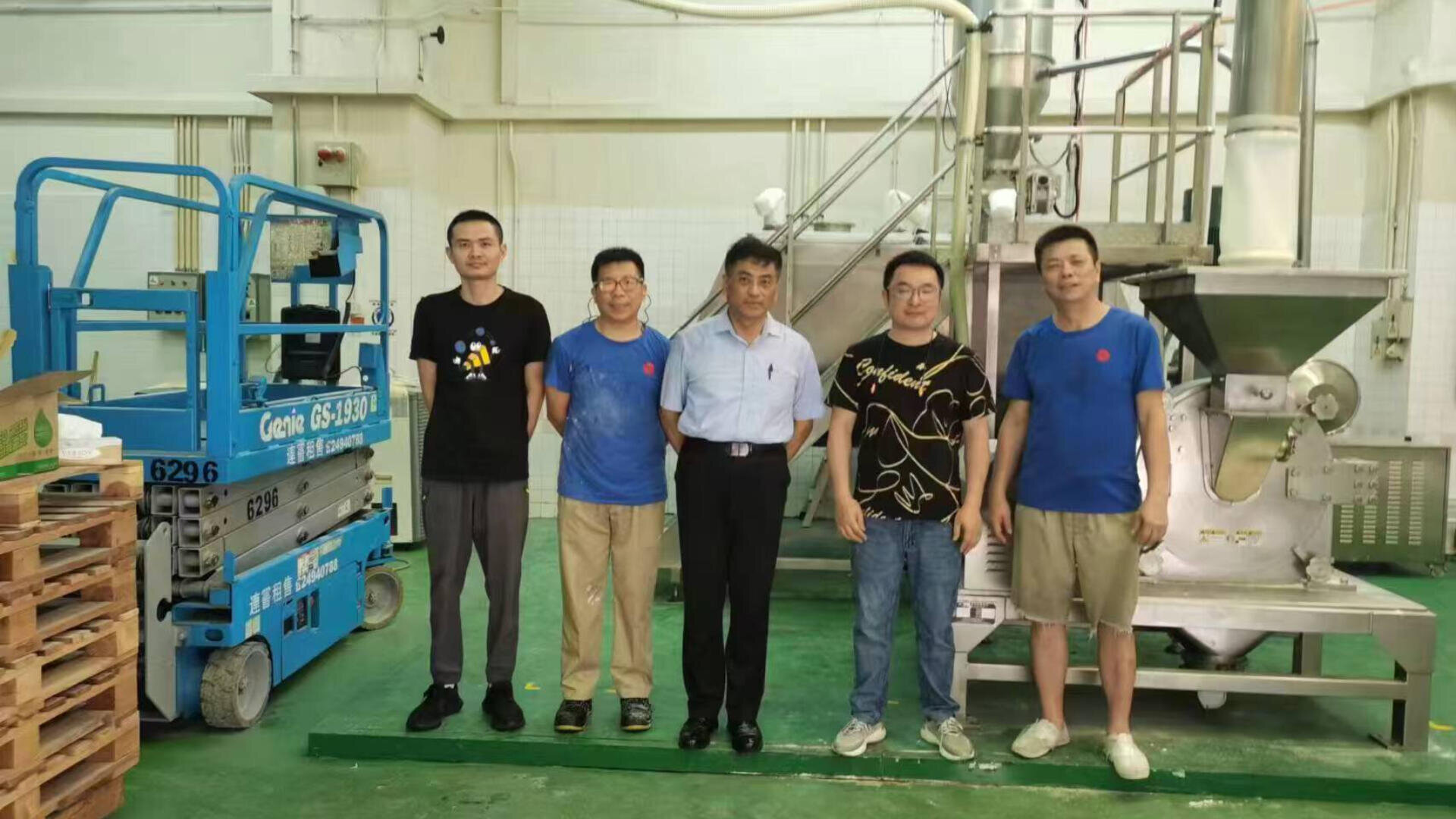 Five men stand side by side in a factory setting next to industrial equipment and a blue lift, posing for a group photo on a green floor with machinery in the background.