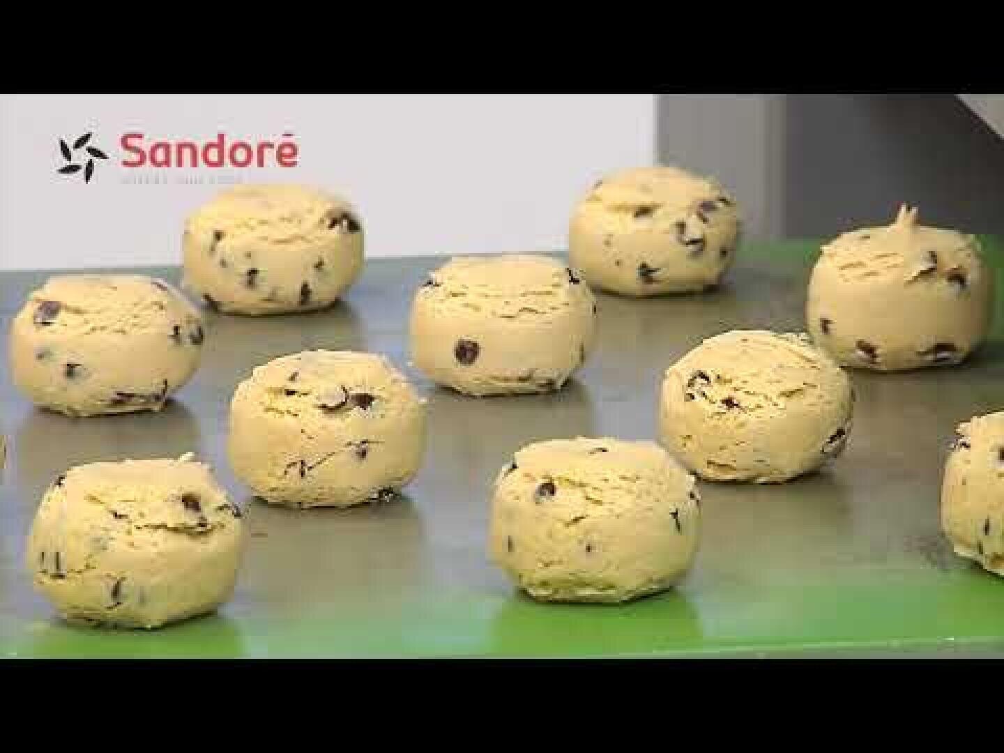 Auf einem grünen Tablett sind mehrere ungebackene, runde Keksteigkugeln aus Schokoladenchips angeordnet. Das Sandoré-Logo ist in der oberen linken Ecke des Bildes zu sehen.