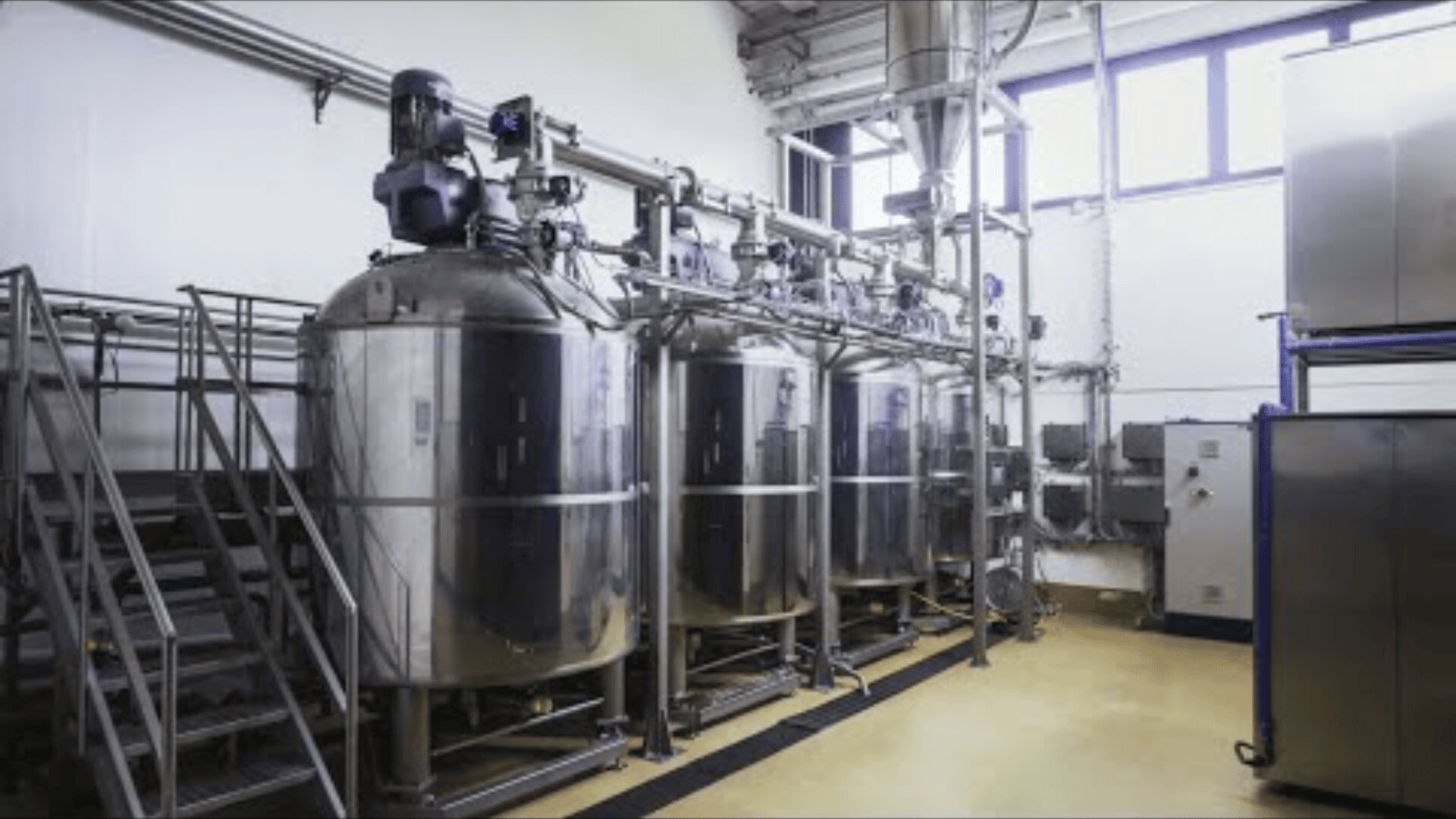 A modern industrial facility with three large stainless steel tanks, metal stairs, and pipes along the walls, used for manufacturing or processing, brightly lit by natural light from high windows.