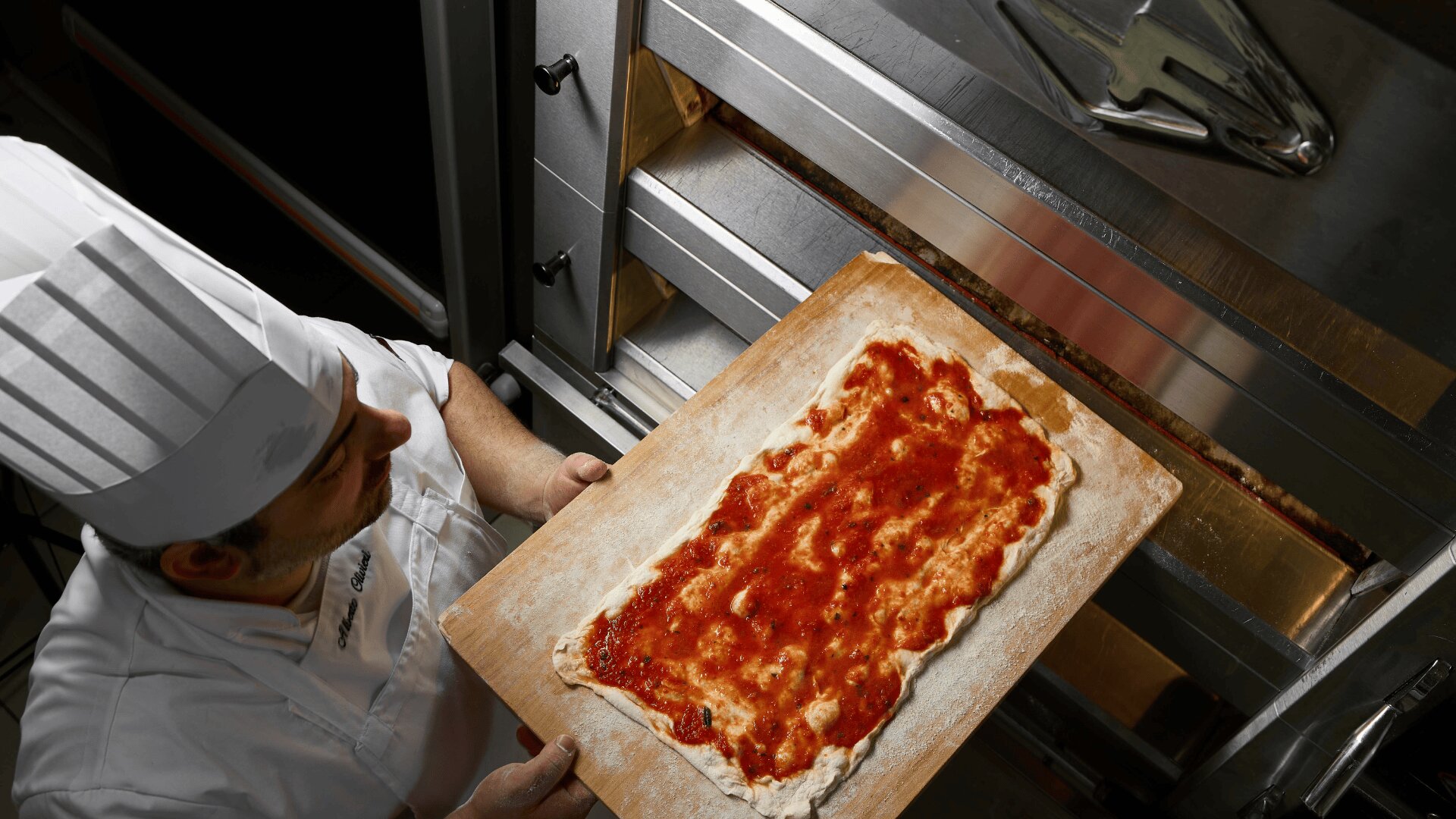 A chef in a white uniform and hat prepares to place a rectangular pizza dough topped with tomato sauce into an open oven using a wooden pizza peel.