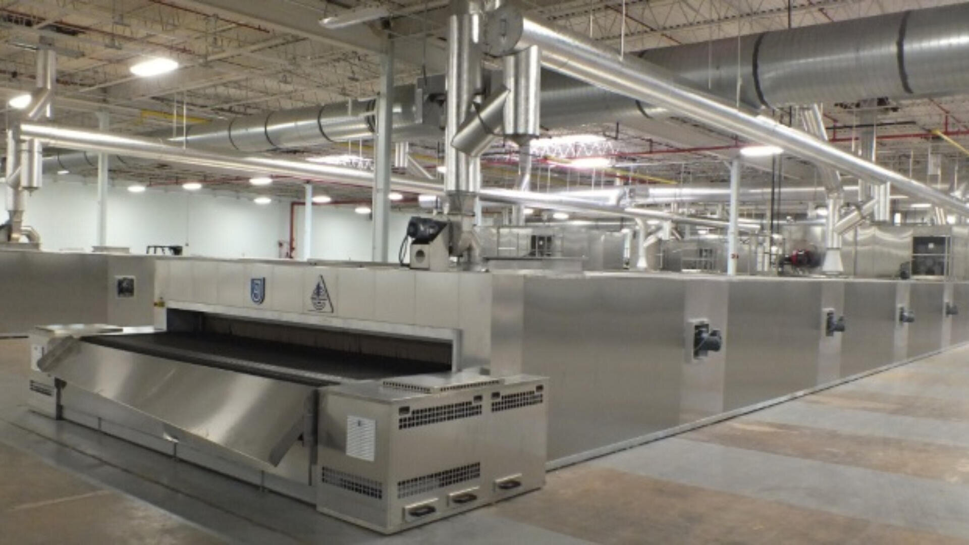 Large industrial conveyor oven in a spacious factory setting, featuring stainless steel surfaces, ventilation ducts above, and a wide conveyor belt for baking or processing food products.