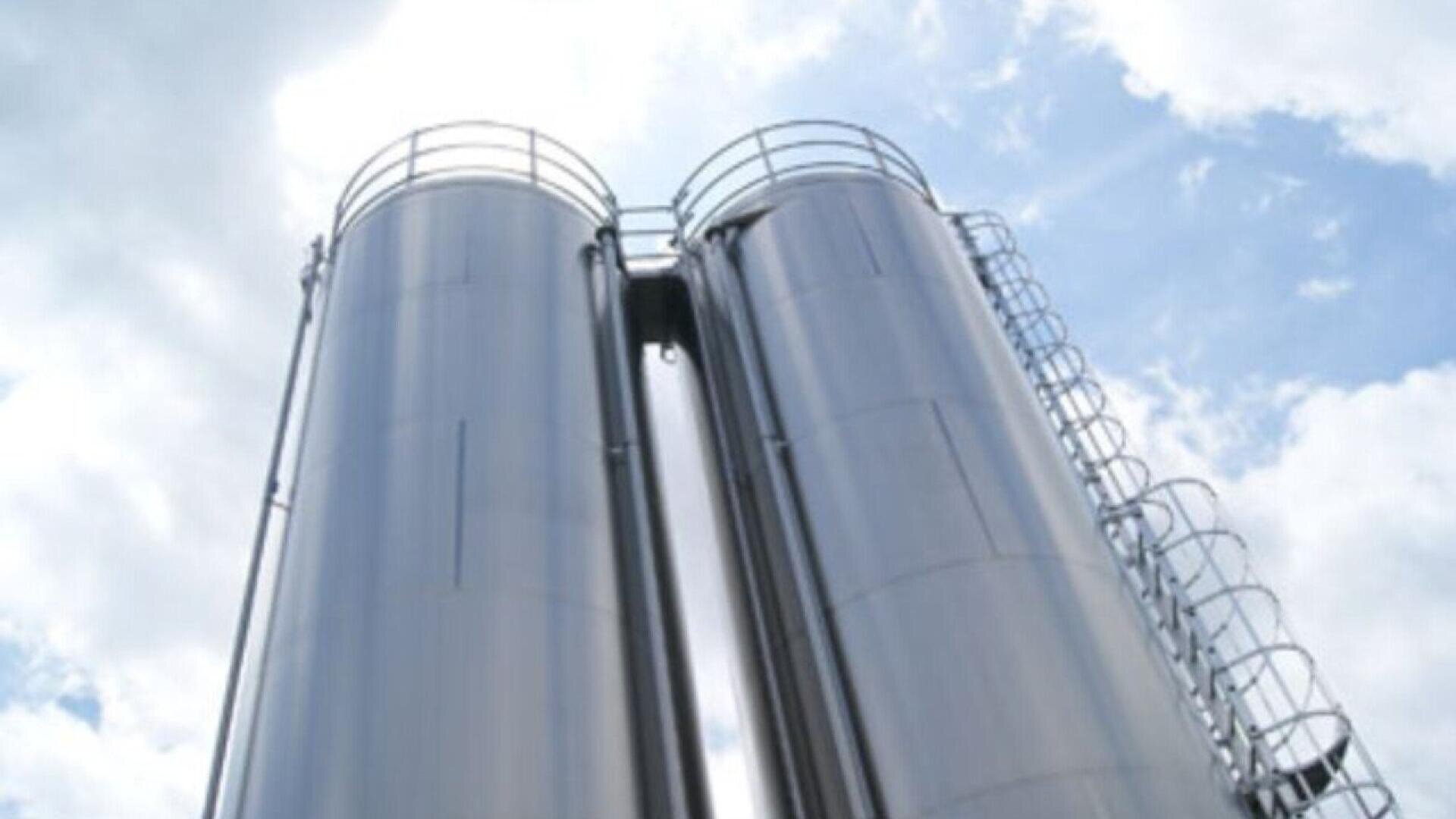 Two tall, shiny metal silos with ladders and railings stand side by side against a partly cloudy sky, viewed from a low angle.