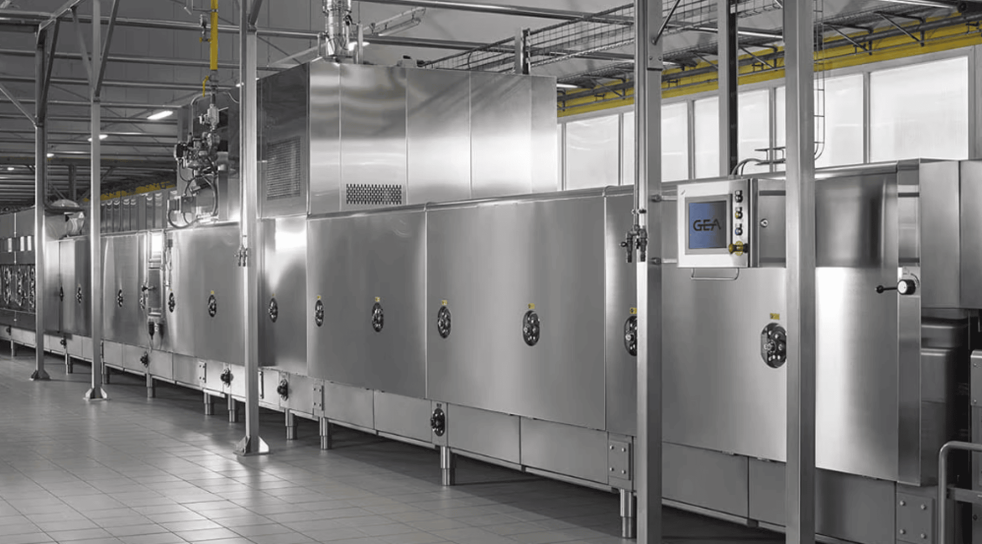 A large stainless steel industrial food processing machine with control panels and piping in a clean, tiled factory environment.