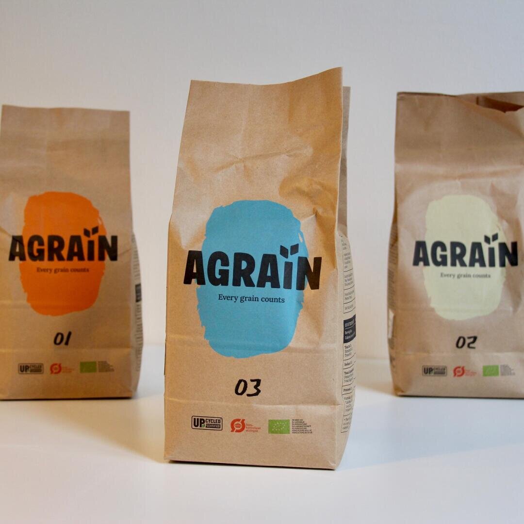 Three brown paper bags labeled AGRAIN Every grain counts stand side by side. Each bag has a different colored label (orange, blue, green) and is numbered 01, 03, and 02 respectively.