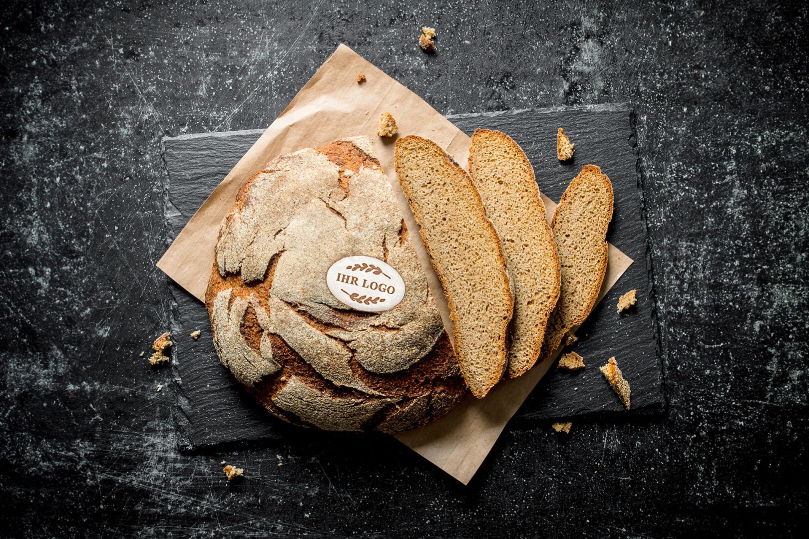 Runder, rustikaler Laib Brot mit dem Etikett „IHR LOGO“ auf der Oberseite, auf braunem Papier und einer dunklen Schiefertafel platziert, mit drei Brotscheiben und Krümeln auf einer strukturierten schwarzen Oberfläche.