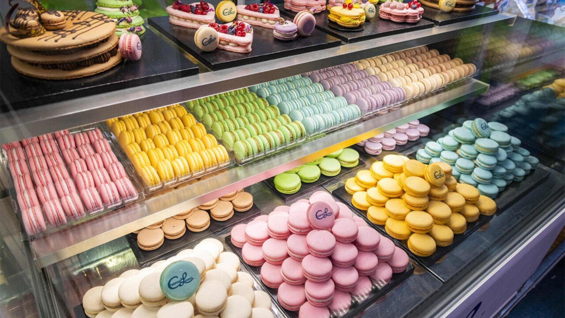 A display case filled with colorful macarons arranged in neat rows, featuring pastel shades of pink, yellow, green, blue, and brown; some have decorative toppings and logos.