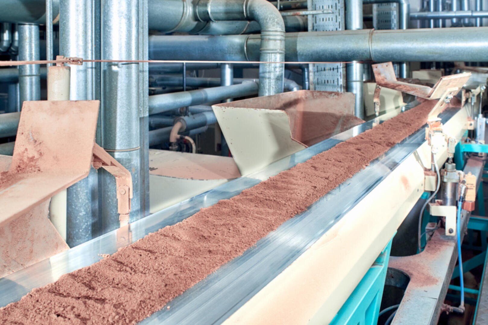 Ein Förderband mit einer kontinuierlichen Schicht aus rötlich-braunem Pulver bewegt sich durch eine Industrieanlage mit Metallrohren und Maschinen im Hintergrund.