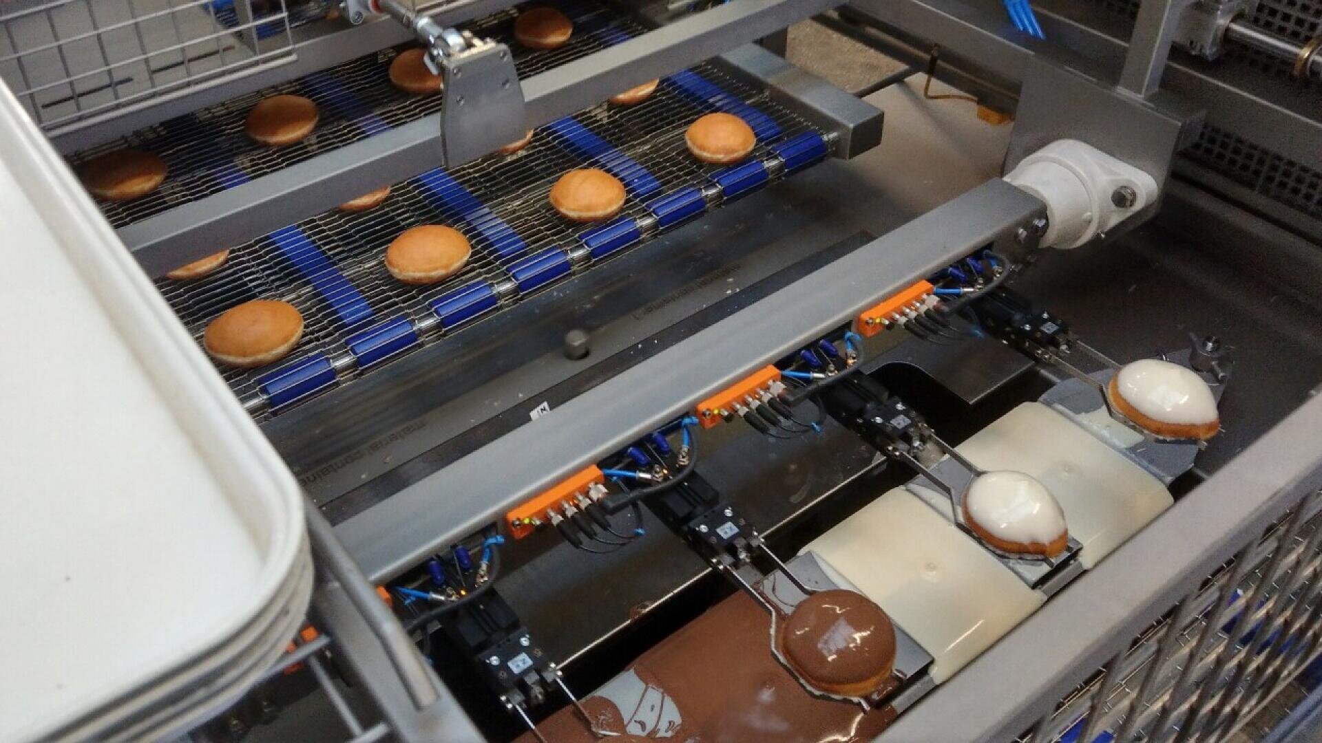 A factory machine dips round pastries into chocolate and white icing, with conveyor belts moving the treats through the automated coating process.