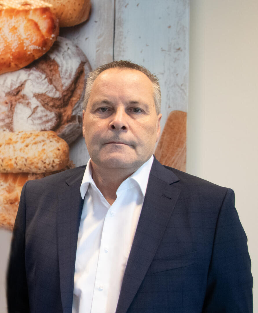 Ein Mann mittleren Alters mit kurzen grauen Haaren, einem dunklen Anzug und einem weißen Hemd steht vor einer Wand, auf der Bilder verschiedener Brotsorten zu sehen sind. Sein Gesichtsausdruck ist ernst, und sein Blick ist direkt in die Kamera gerichtet.