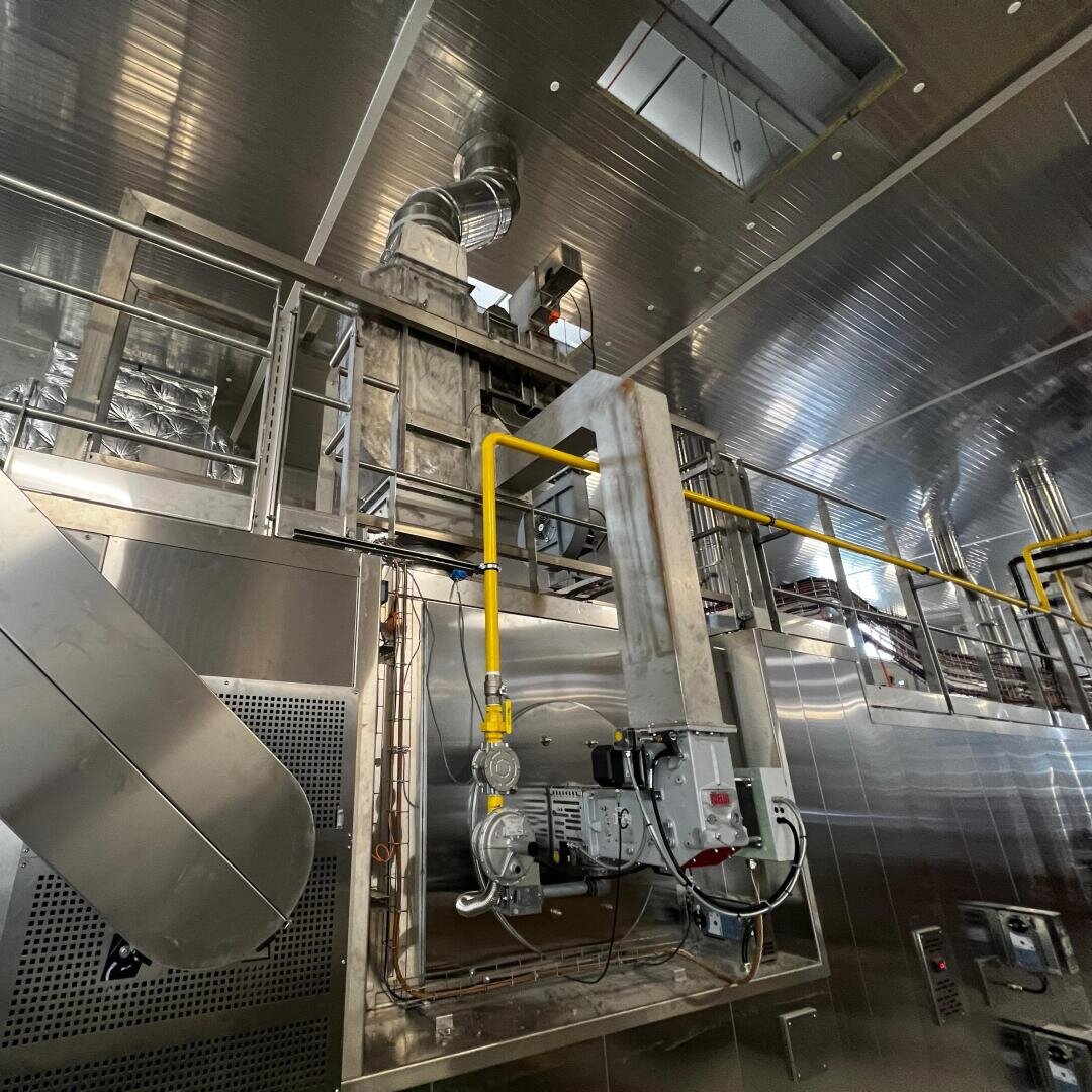 Large industrial facility interior with metallic walls, pipes, ducts, and machinery. Yellow gas piping and control panels are visible, along with elevated walkways and vents on the ceiling. The environment looks clean and modern.