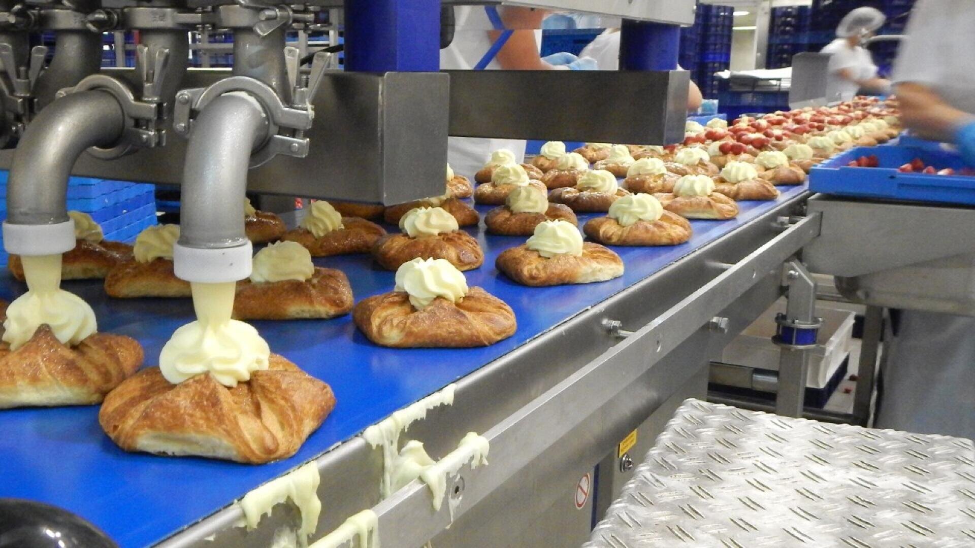 Pastries on a conveyor belt being topped with cream by a machine in a bakery or food production facility, with workers in the background handling trays.