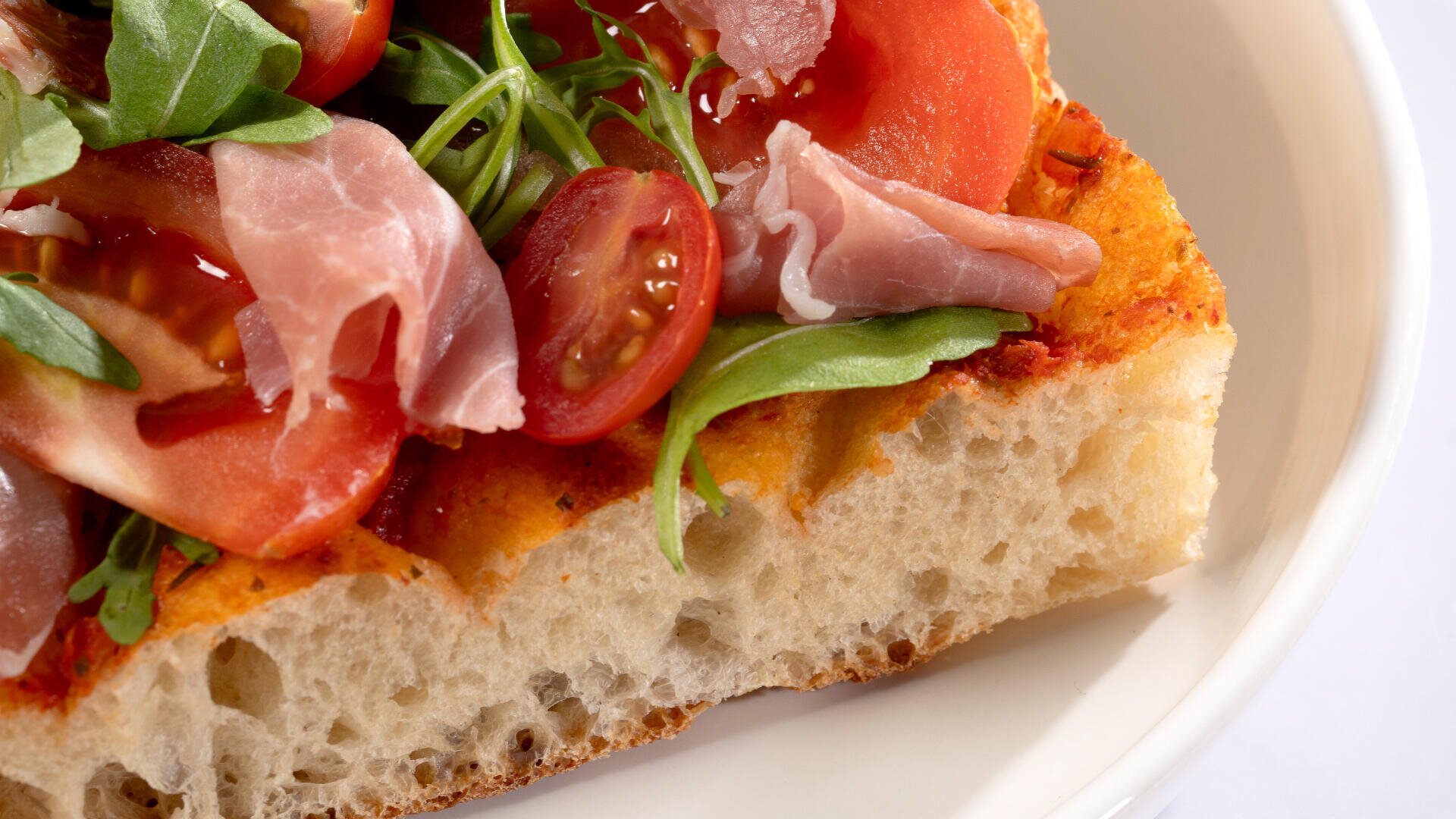 A close-up of a slice of focaccia bread topped with arugula, cherry tomatoes, prosciutto, and tomato slices on a white plate.