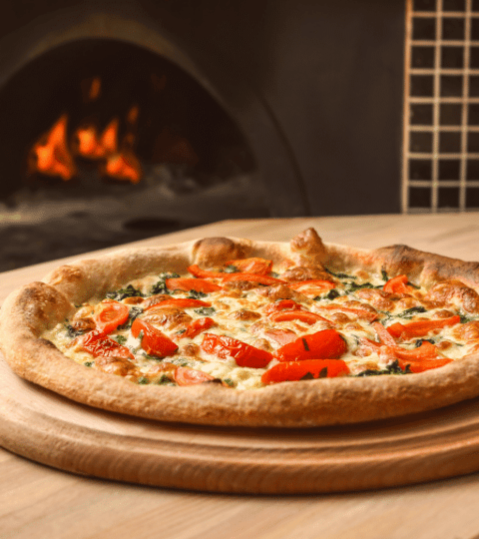 A freshly baked pizza topped with tomatoes, cheese and herbs lies on a wooden board in front of a traditional wood-fired oven with the flames visible.