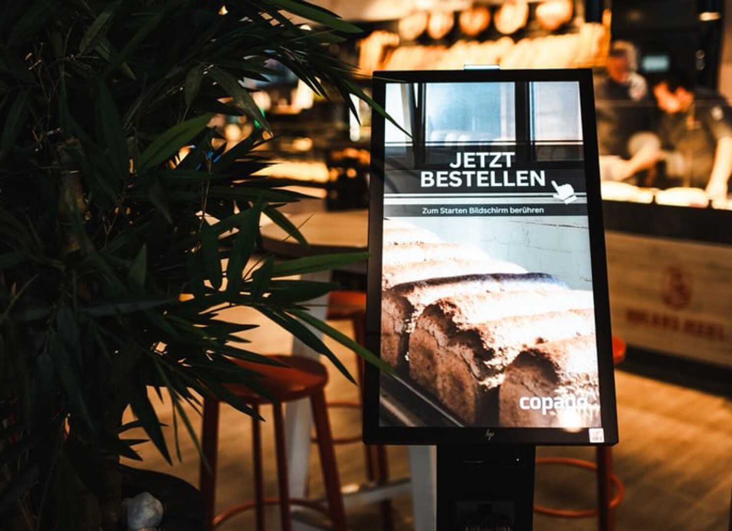 A touchscreen kiosk in a bakery displays a German sign reading JETZT BESTELLEN (Order Now) with an image of bread. The background shows blurred bakery shelves and seating, with a leafy plant in the foreground.