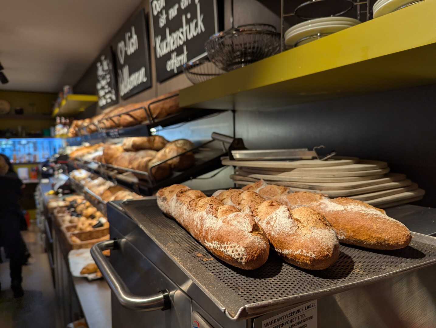 Eine Bäckereitheke präsentiert frische Baguettes und verschiedene Brotsorten auf Tabletts und Regalen. Darüber hängen Speisekarten auf Kreidetafeln mit deutscher Aufschrift. Teller und Körbe sind in der Nähe gestapelt. Im Hintergrund sind weitere Backwaren und ein sanft beleuchteter Innenraum zu sehen.