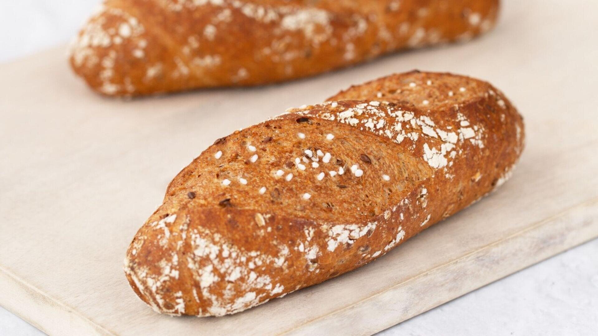 A close-up of two loaves of rustic whole grain bread with a golden brown crust and sprinkled oats, resting on a light wooden surface.