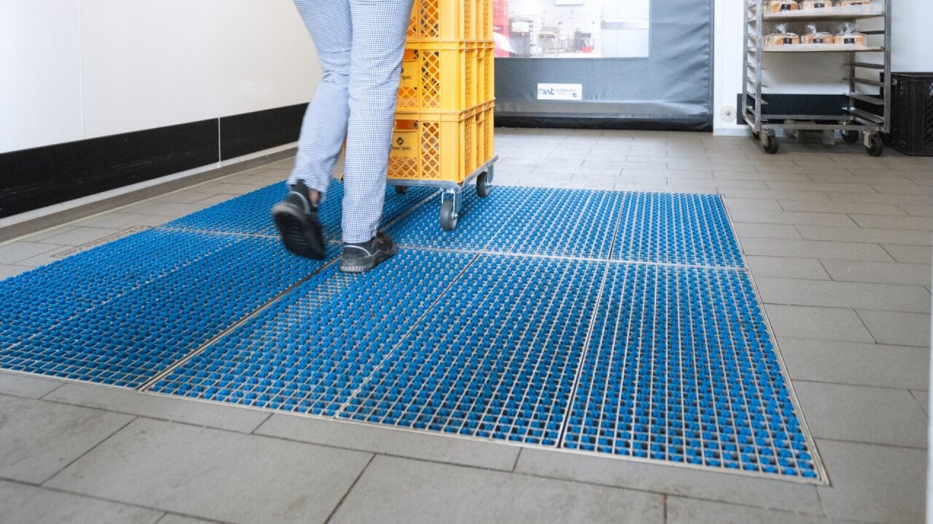 Person wearing checkered pants and black shoes pushes a cart with yellow crates over a blue sanitizing mat with metal grid in an indoor, tiled-floor setting.