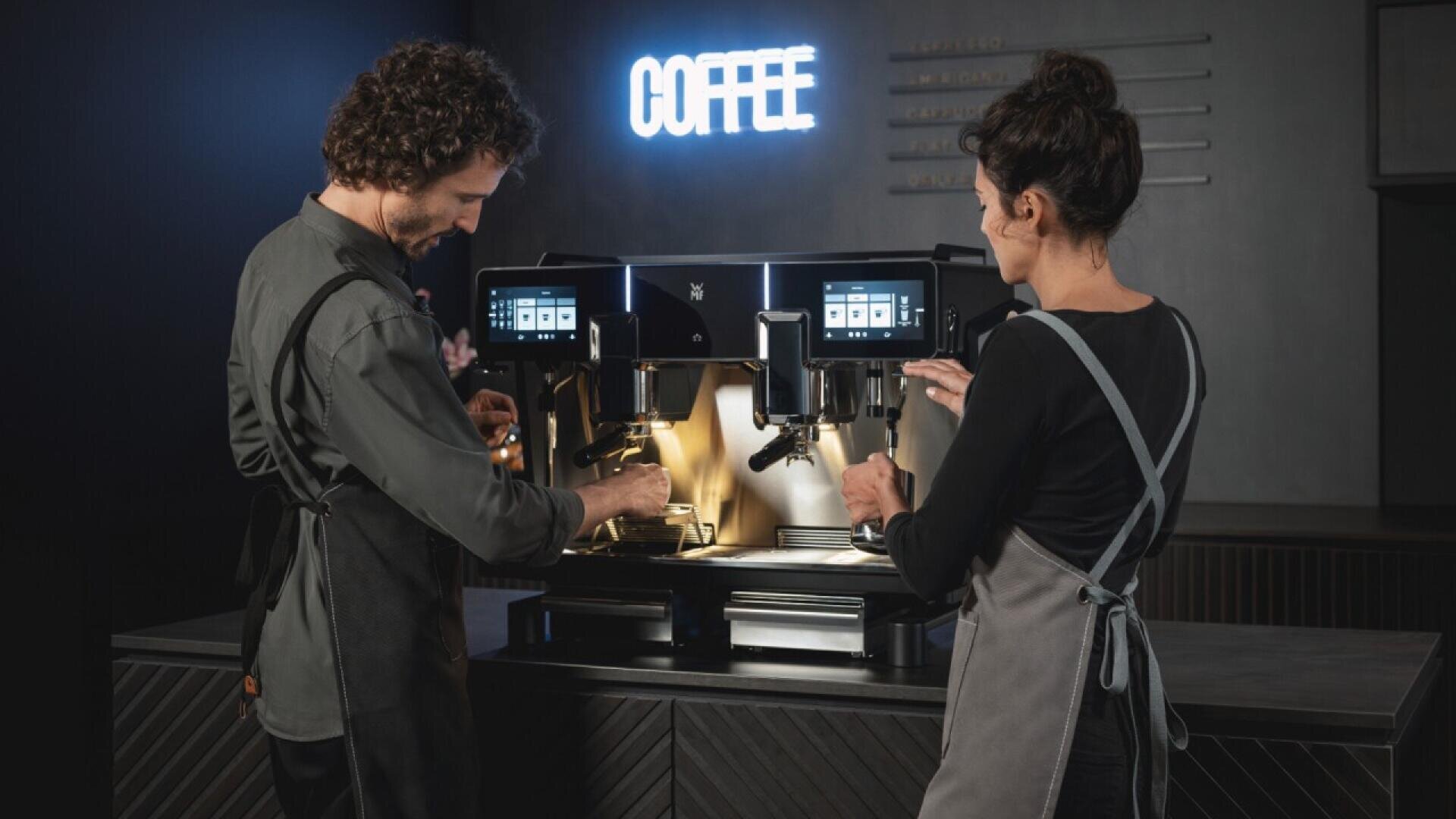 Two baristas in aprons operate a large, modern espresso machine in a dimly lit café. A neon “COFFEE” sign glows on the wall behind them.