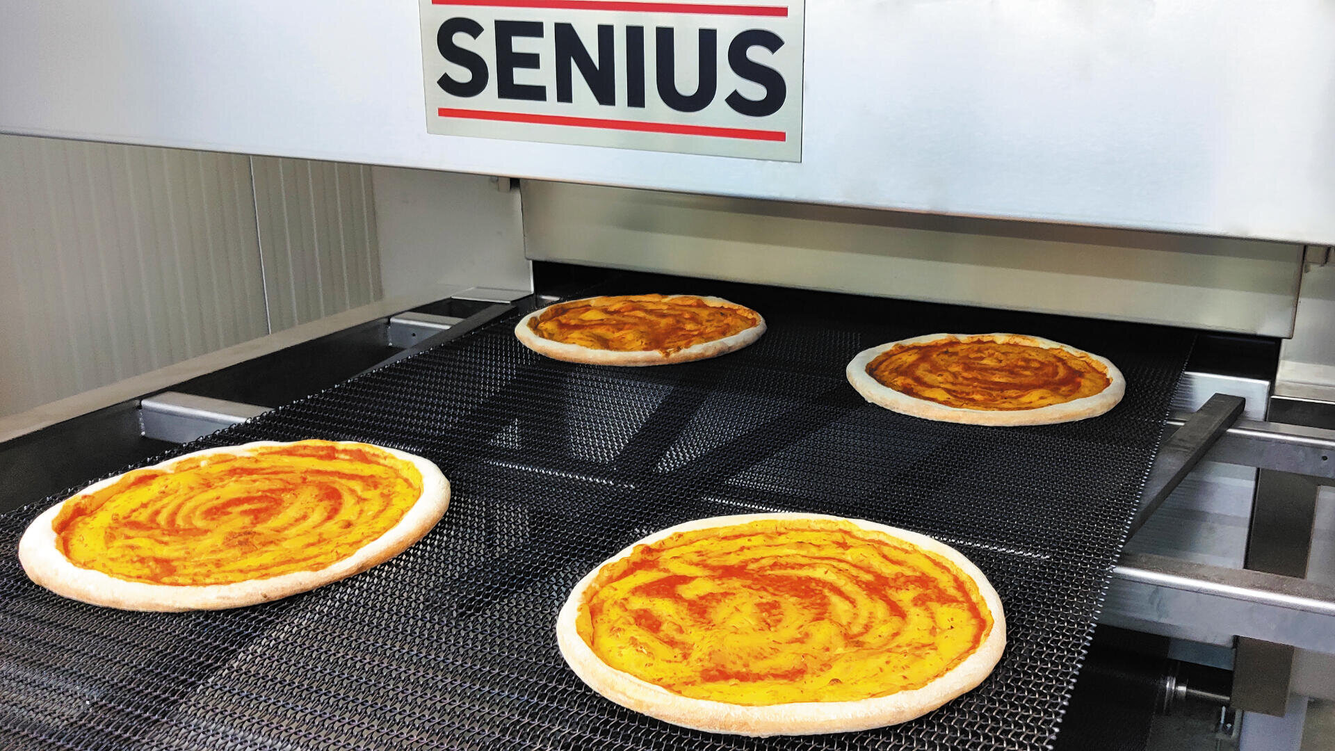 Vier rohe Pizzaböden mit Tomatensauce laufen auf einem Förderband unter einer Maschine mit der Aufschrift SENIUS, die wahrscheinlich Teil eines automatisierten Pizzaproduktionsprozesses ist.