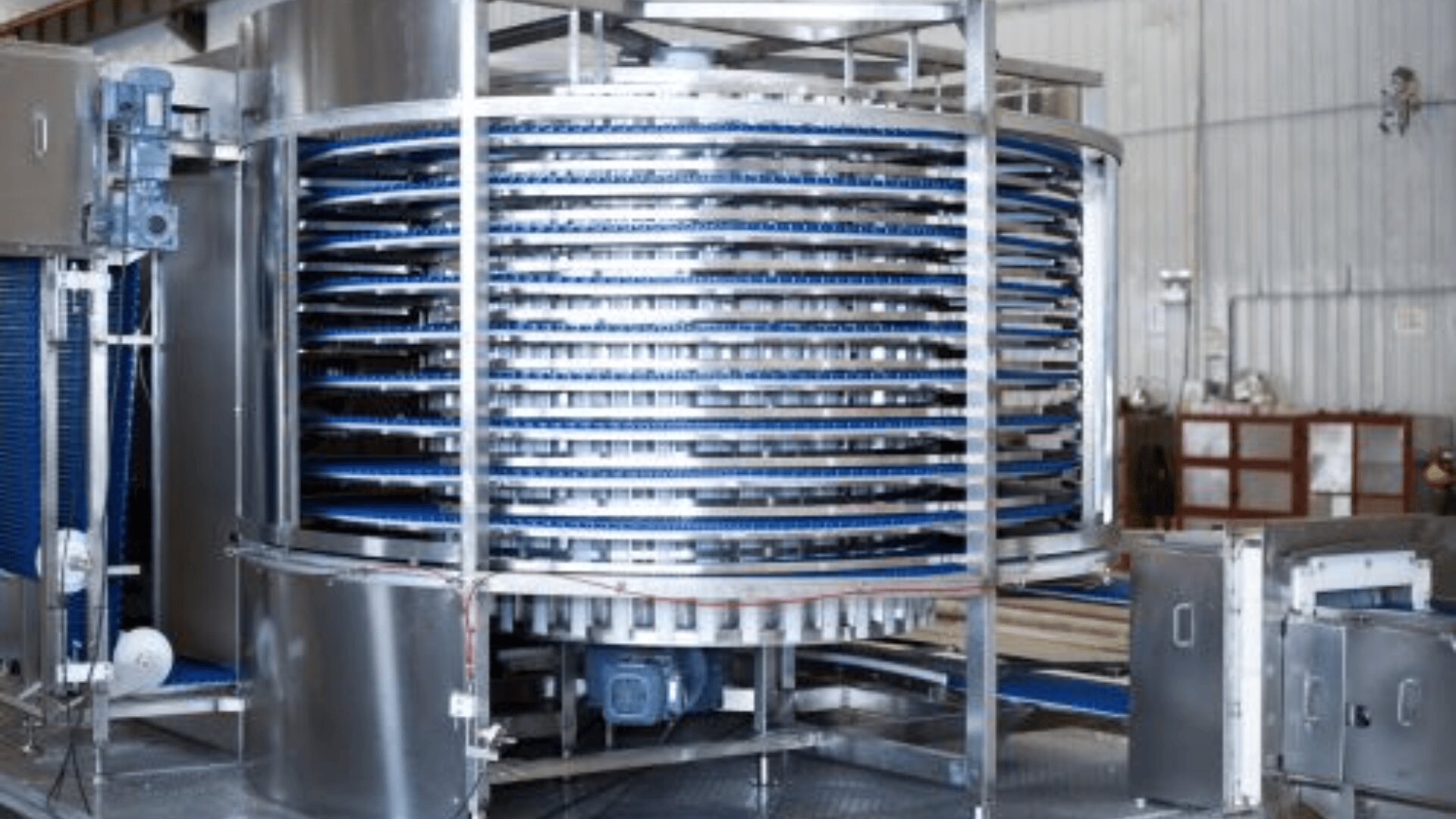 A large industrial spiral conveyor system made of stainless steel with multiple blue conveyor belts stacked in a circular tower inside a factory or warehouse setting.