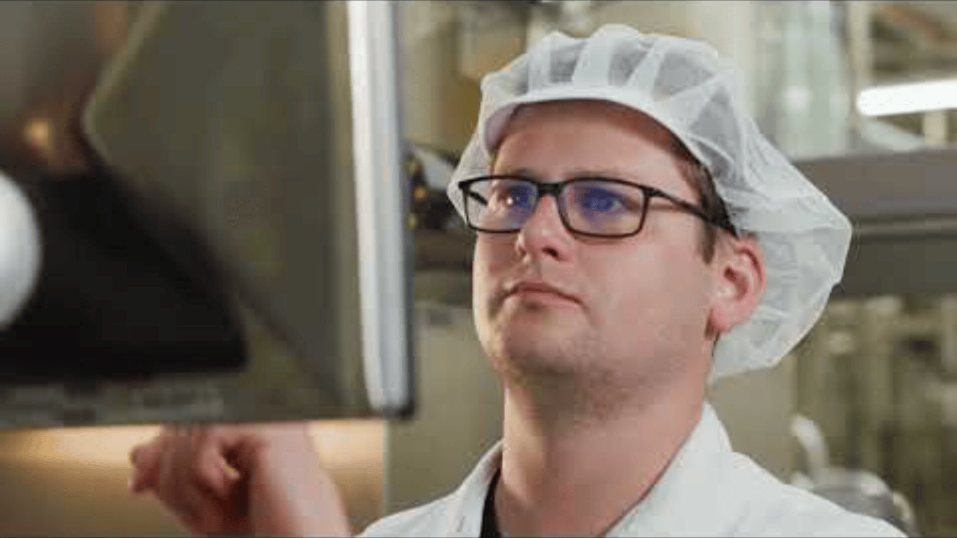 Eine Person, die eine Brille, ein weißes Haarnetz und einen Laborkittel trägt, konzentriert sich auf die Bedienung eines Touchscreens in einer Labor- oder Industrieumgebung.