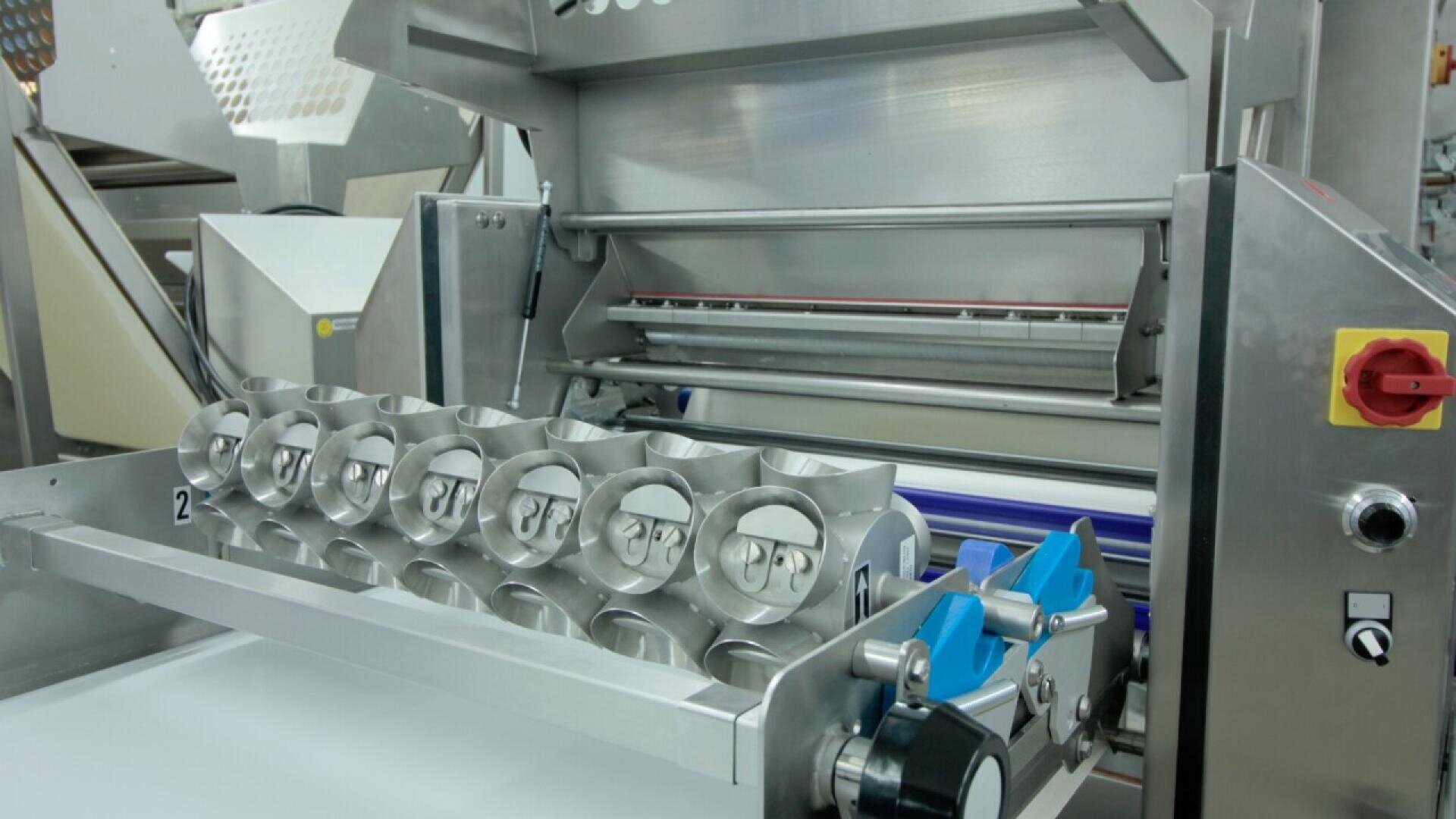 Close-up of an industrial food processing machine with metal rollers, blades, and various mechanical parts, used for shaping or cutting dough in a factory setting.