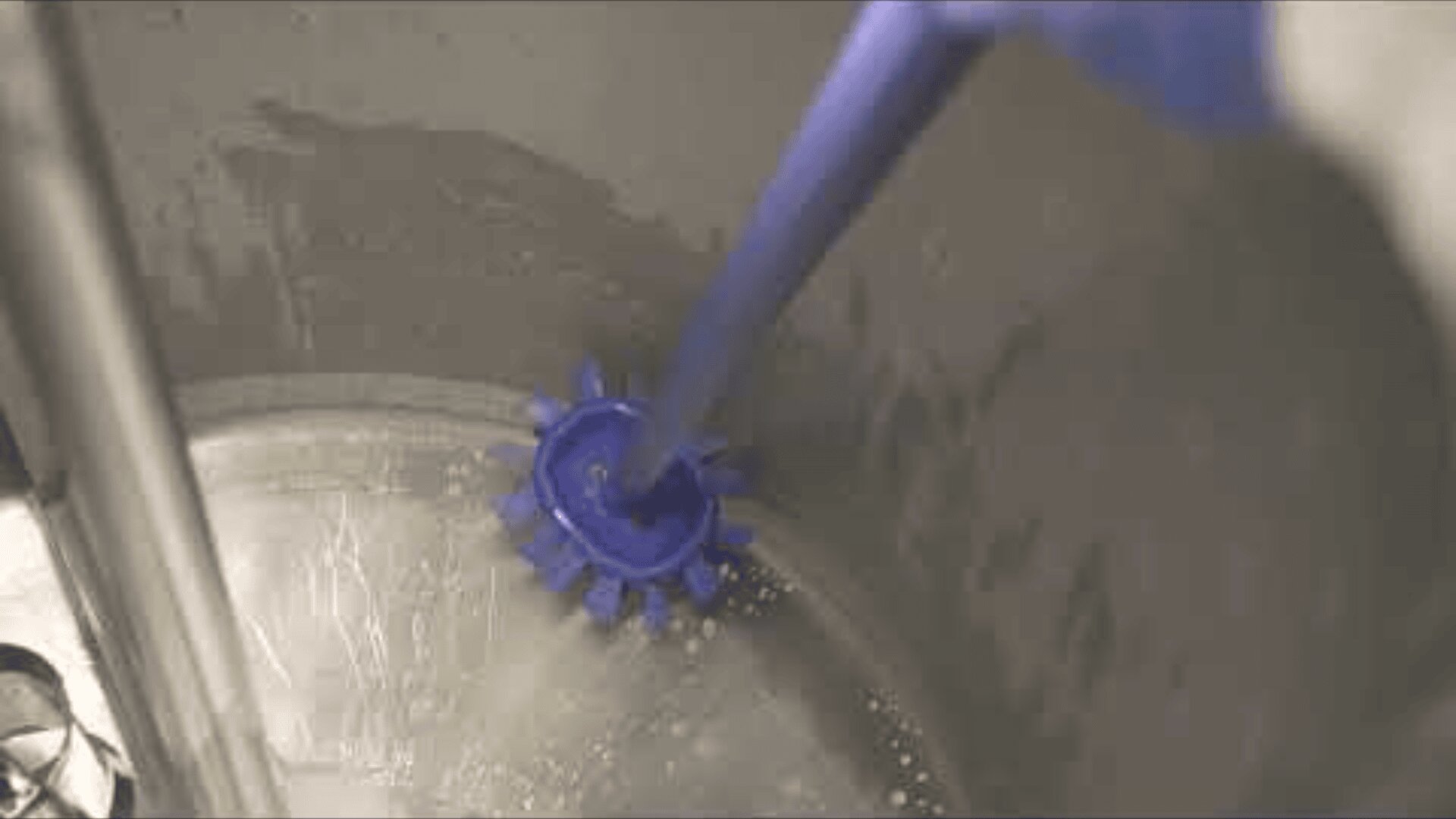 A close-up of a purple cleaning brush being used to scrub the inside of a stainless steel sink or container, with some water and soap suds visible on the surface.