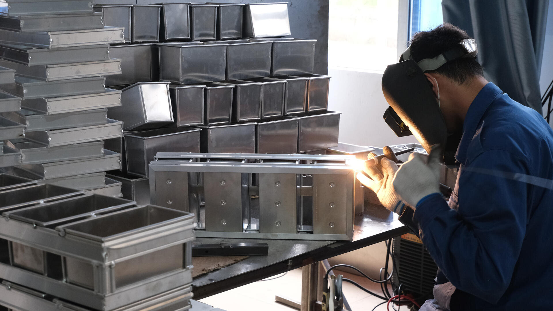 A person wearing protective gear welds metal parts at a workbench, surrounded by stacks of shiny metal containers in a workshop.