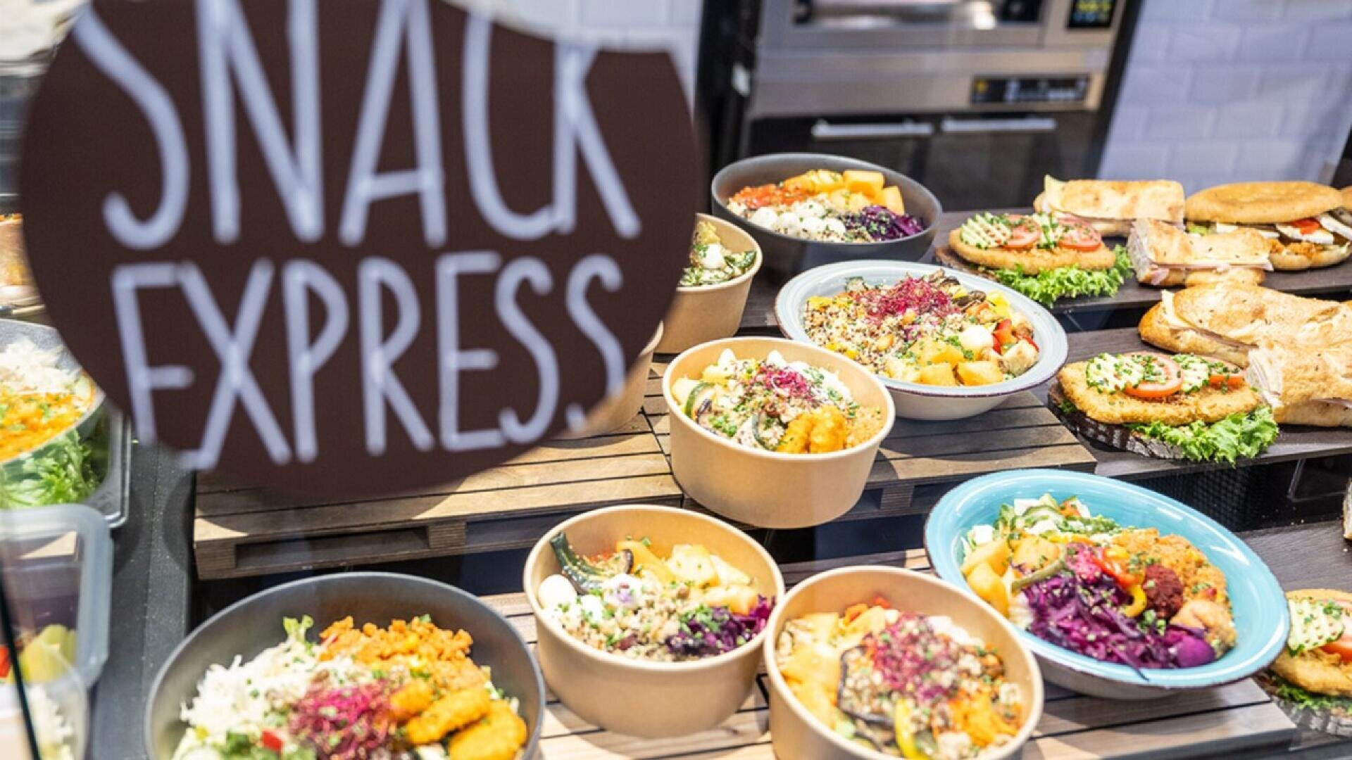 A display of assorted colorful salads and sandwiches in bowls and plates at a deli counter, with a sign reading SNACK EXPRESS in the foreground.