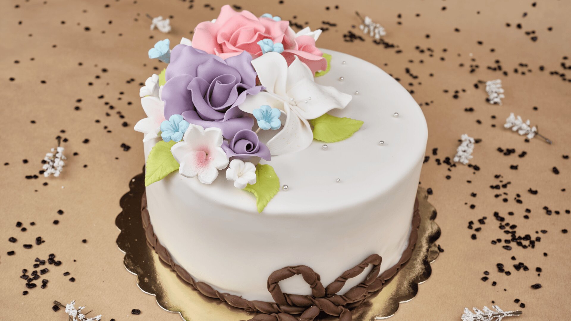 A round white cake decorated with pastel fondant flowers, including purple, pink, and white blooms with green leaves. The cake sits on a gold board, surrounded by scattered chocolate sprinkles.