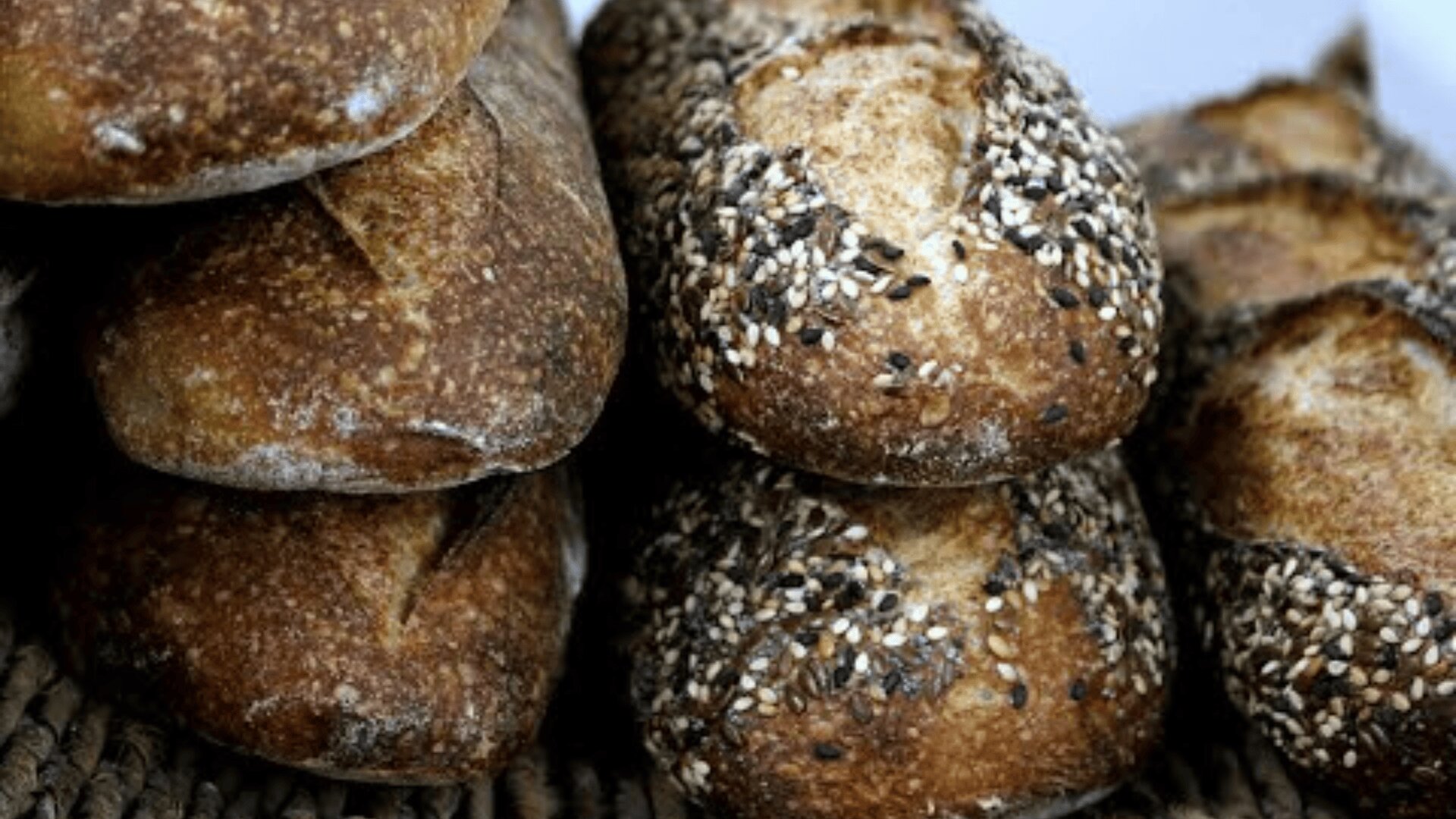 Several rustic loaves of bread are stacked together; some have a golden crust with crackled surfaces, while others are topped with a mix of seeds, including sesame and sunflower seeds.