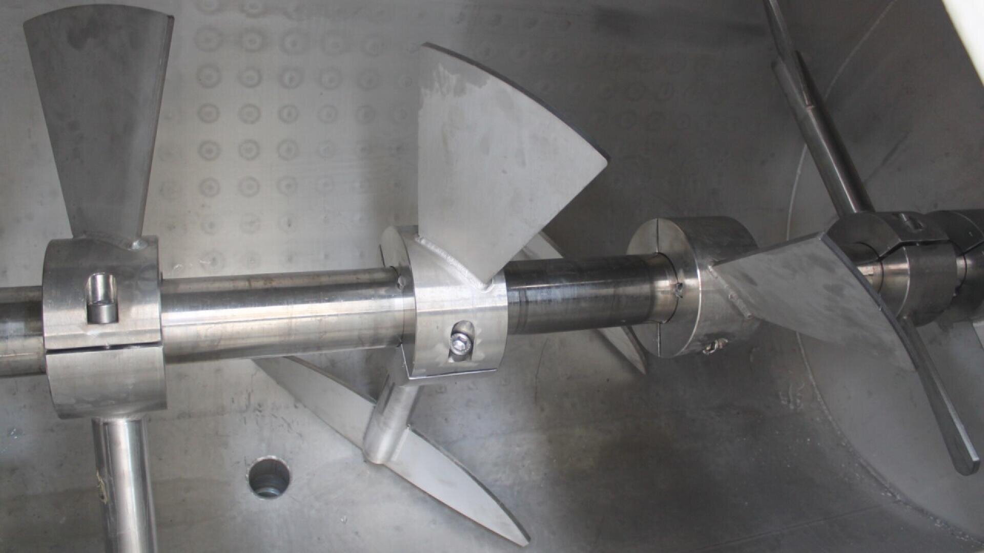 Close-up view of a stainless steel industrial mixer with multiple angled blades attached to a central rotating shaft inside a metal chamber.