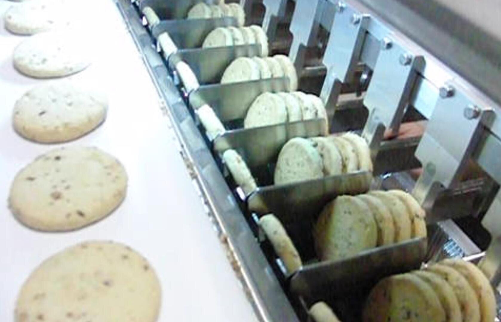 Rows of round cookies with chocolate chips are being sorted and aligned by a metal machine on a conveyor belt in a food processing facility.