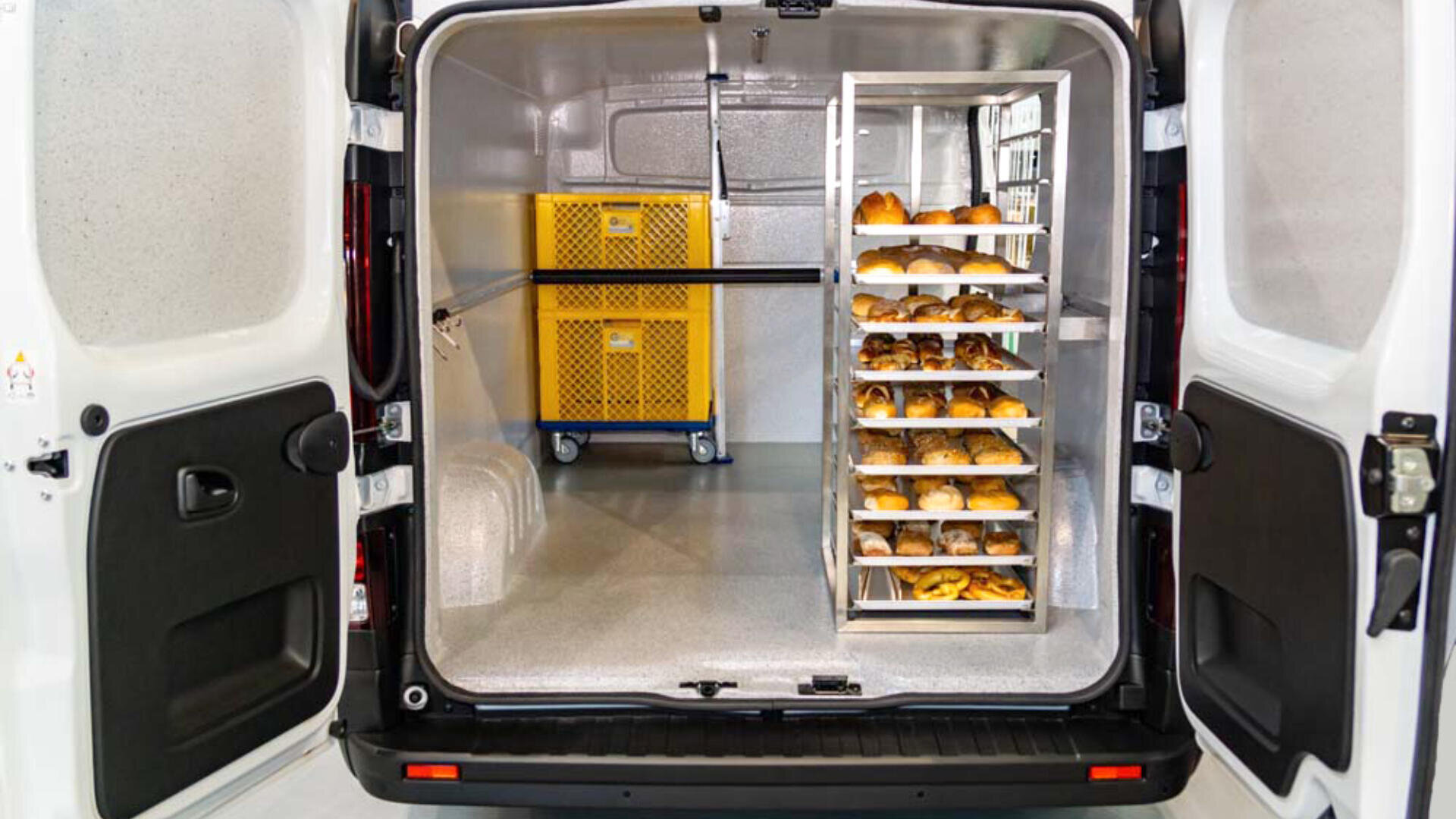 Die offene Rückseite eines Lieferwagens zeigt Tabletts mit verschiedenen Brötchen auf einem Metallgestell und einen gelben Wagen mit Behältern auf einer sauberen, gut beleuchteten Ladefläche.