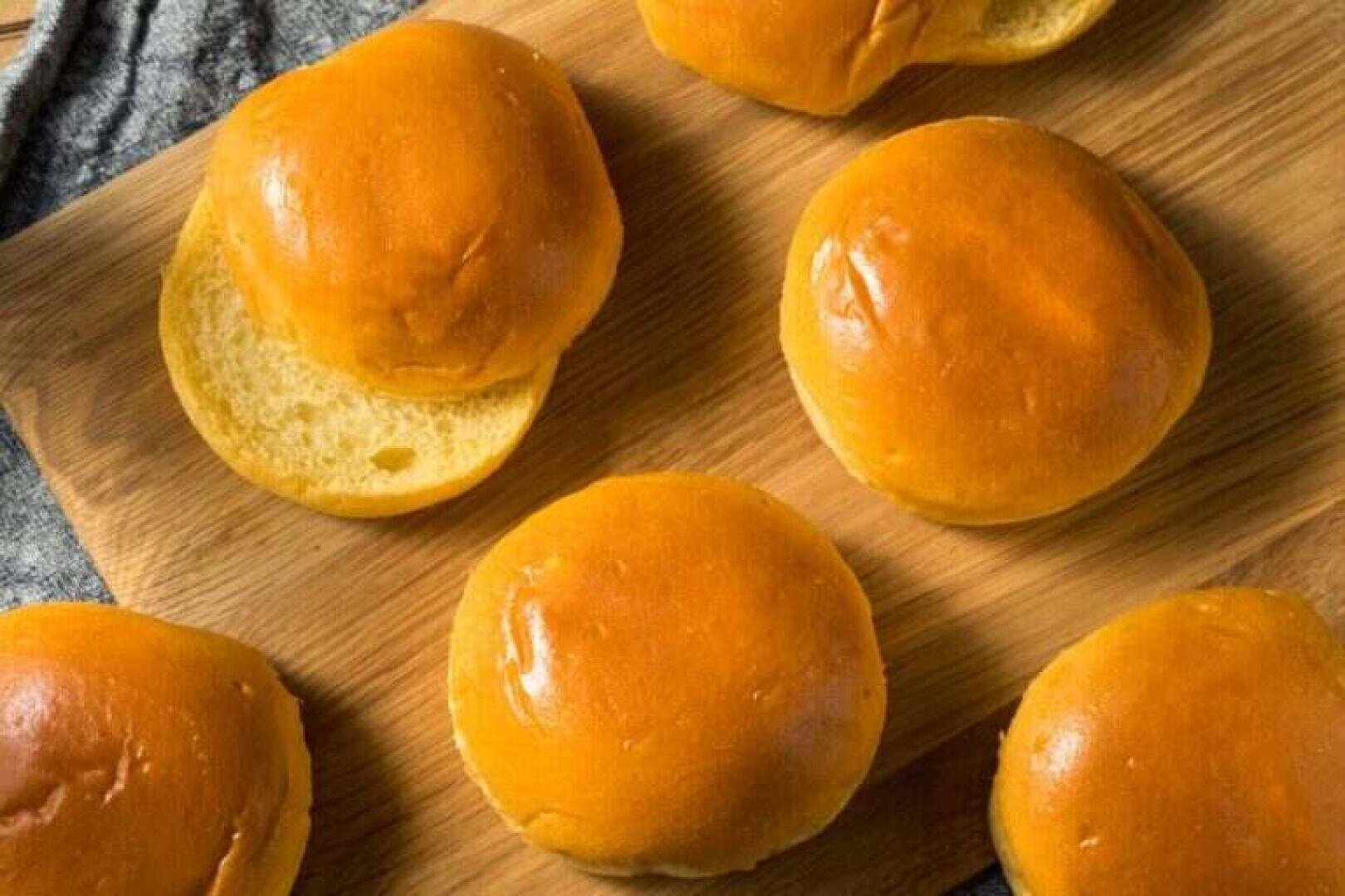 Sechs goldbraune Brioche-Brötchen sind auf einer Holzunterlage angeordnet, wobei ein Brötchen teilweise geöffnet ist, um sein weiches, fluffiges Inneres zu zeigen. Die Oberfläche und die Textur der Brötchen wirken glänzend und frisch.
