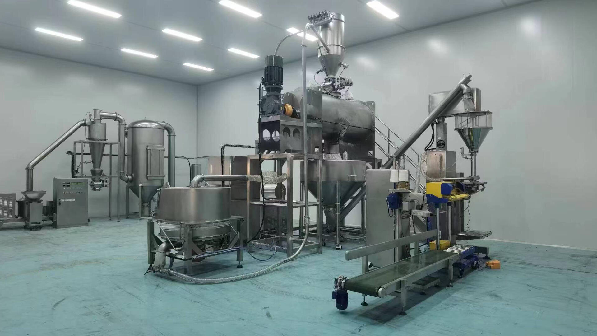 A modern industrial food processing machine setup in a clean, spacious room with stainless steel tanks, pipes, conveyors, and automated packaging equipment under bright overhead lighting.