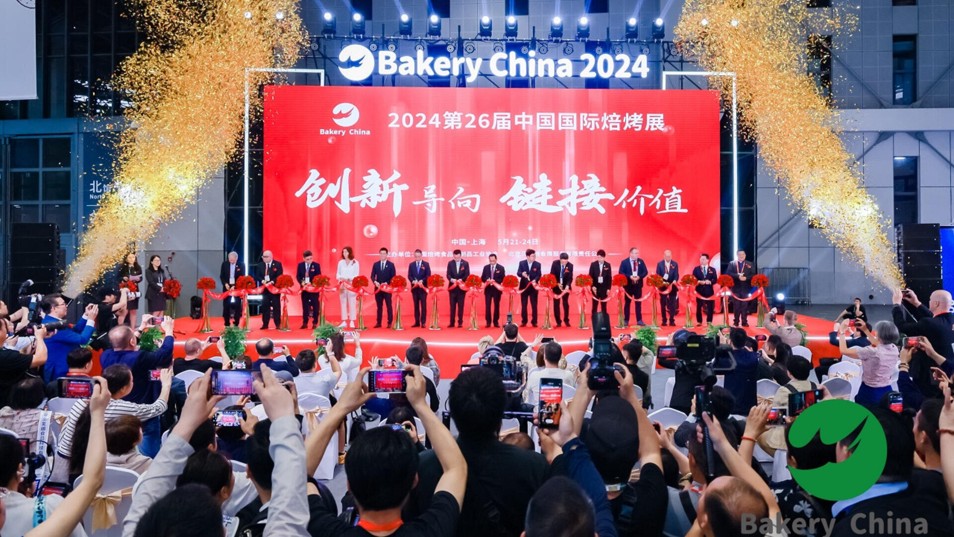 A large audience photographs a ribbon-cutting ceremony on stage at Bakery China 2024. Event staff and officials stand in a line with red ribbons, under banners and bright lights, in a decorated exhibition hall.