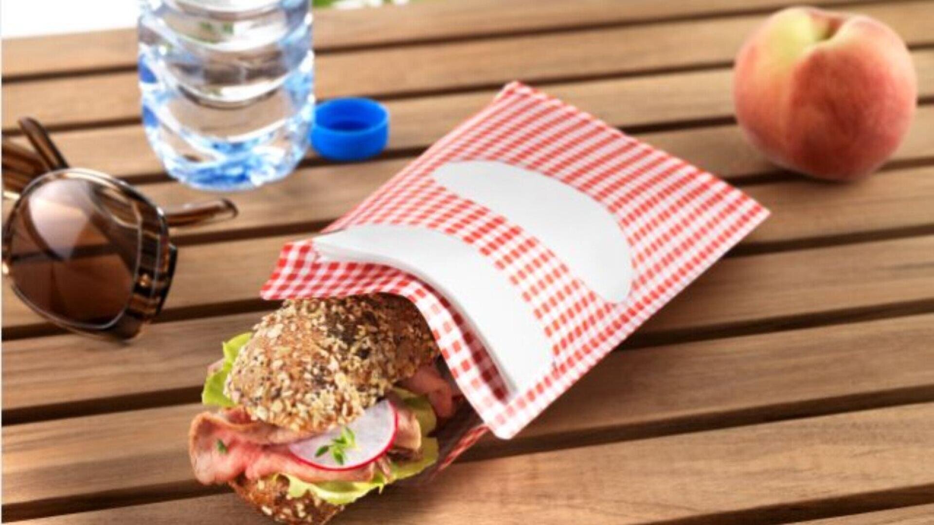 Ein Mehrkornsandwich mit Salat, Schinken und Rettich liegt in einer rot karierten Papierhülle auf einem Holztisch neben einem Pfirsich, einer Flasche Wasser und einer Sonnenbrille.