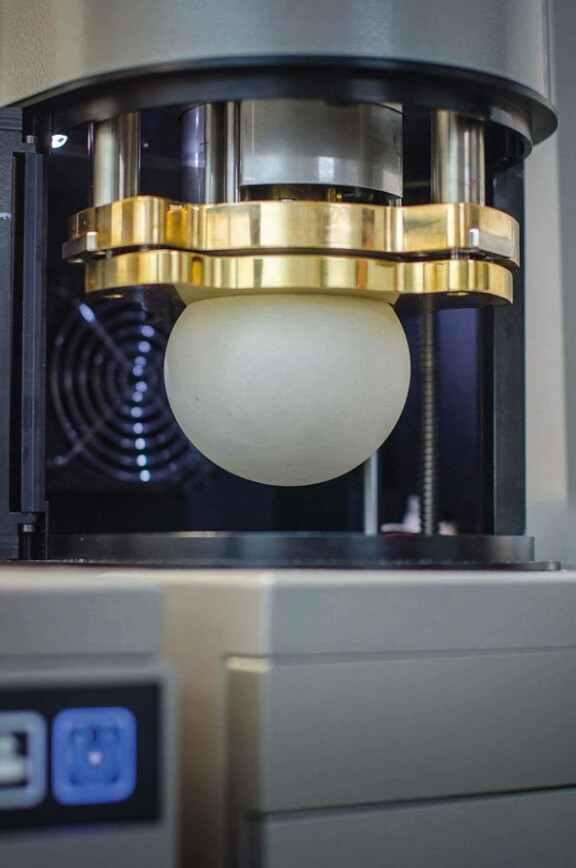 Eine glatte, runde Teigkugel wird in einer Metallmaschine von einer goldfarbenen Platte gepresst, wahrscheinlich zum Testen oder Formen. Im Hintergrund ist ein schwarzer Fächer zu sehen.