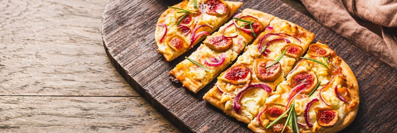 Eine rechteckige Fladenbrotpizza mit Feigen, roten Zwiebelscheiben, Käse und Kräutern liegt auf einem rustikalen Holzbrett, wobei ein Stück leicht vom Rest abgesetzt ist.
